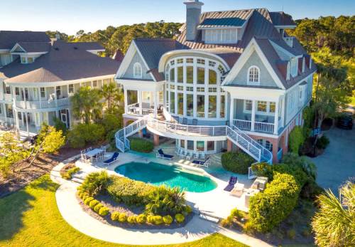 Aerial view of a large, elegant house with a pool, surrounded by lush landscaping and other homes in the background.