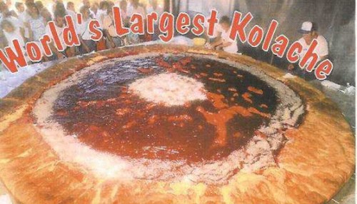 A massive kolache topped with various fillings, surrounded by people, with "World's Largest Kolache" text above.