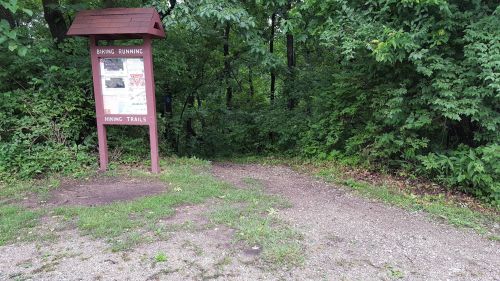 A trailhead sign for biking and running, surrounded by lush green trees and a gravel path leading into the woods.