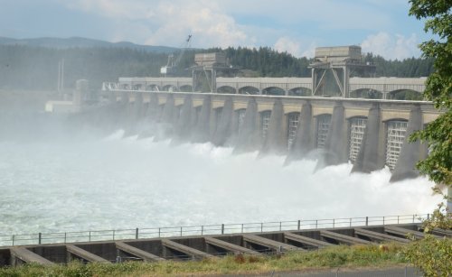 A large dam releasing water, creating a misty spray, with a backdrop of trees and a cloudy sky.
