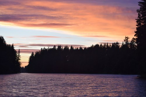 A serene river at sunset, with vibrant orange and purple skies and silhouettes of trees lining the water's edge.
