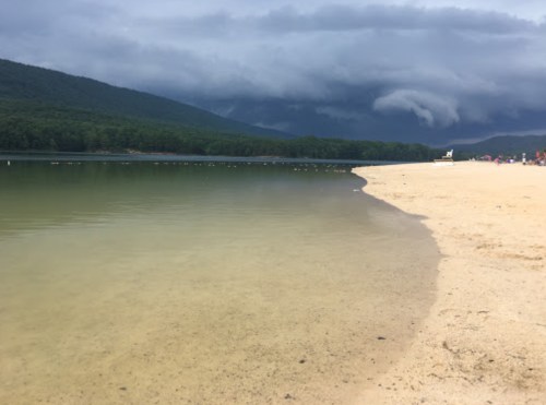 A calm lake with a sandy shore, surrounded by green hills and dark, cloudy skies in the background.