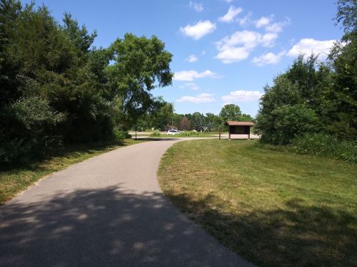 A winding path through greenery leads to a grassy area with a shelter and blue sky in the background.