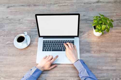 A person typing on a laptop with a coffee cup and a small plant on a wooden table. The laptop screen is blank.