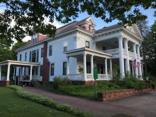 A large, historic white house with columns, a porch, and an American flag, surrounded by greenery and trees.