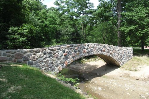 A stone arch bridge spans a dry creek, surrounded by lush green trees and grass in a serene park setting.