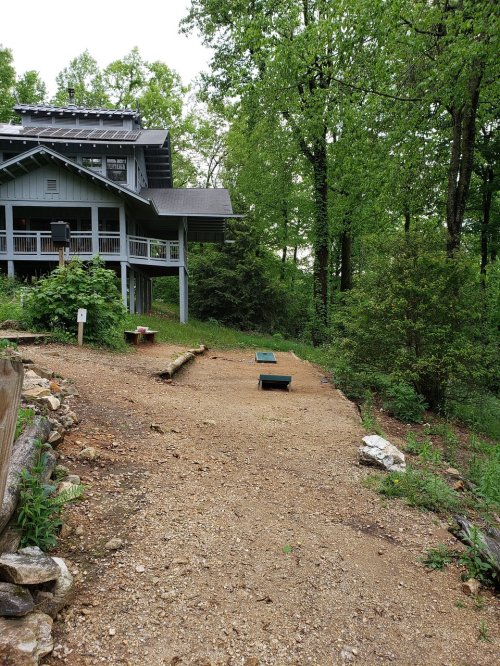 A gravel path leads to two green bean bag toss boards, surrounded by trees and a house in the background.