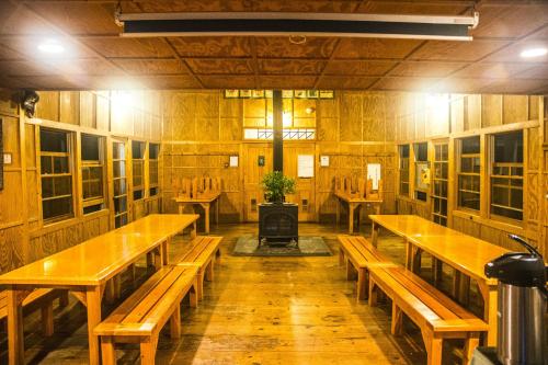 A cozy wooden dining area with long tables, benches, and a central stove, illuminated by warm lighting.
