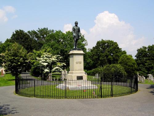 A statue of a soldier stands in a fenced area surrounded by trees and grass under a blue sky with clouds.