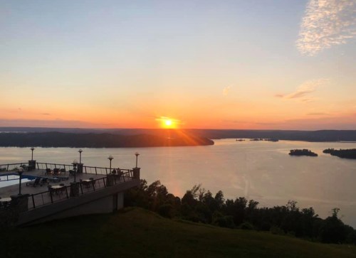 Sunset over a tranquil lake, with silhouettes of trees and a balcony in the foreground, casting a warm glow.