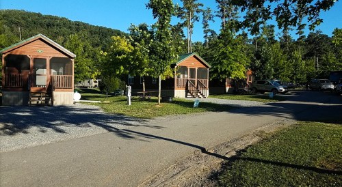 Two wooden cabins with porches are set among trees in a gravel campground, with a road running through the area.
