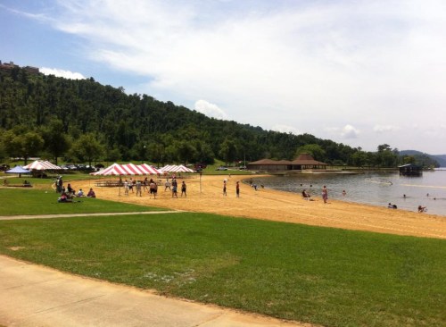 A sandy beach with volleyball nets, tents, and people enjoying the sun by a lake surrounded by green hills.