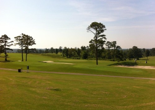 A scenic view of a golf course with lush green grass, trees, and a sand trap under a clear blue sky.