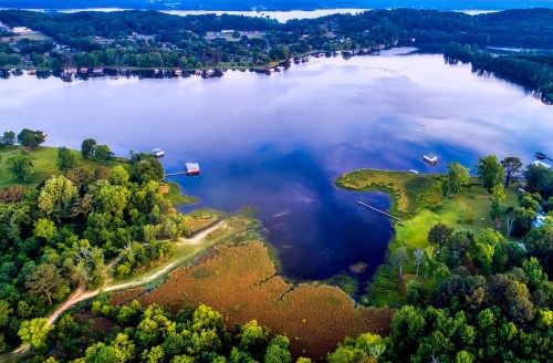 Aerial view of a serene lake surrounded by lush greenery and small boats, with a peaceful shoreline and distant hills.