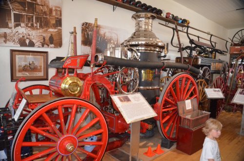 A vintage fire engine displayed in a museum, featuring red wheels and shiny metal components, with a child nearby.