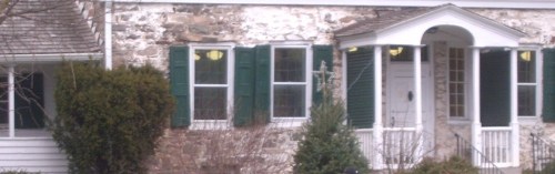 A historic stone house with green shutters and a white porch, surrounded by shrubs and trees.