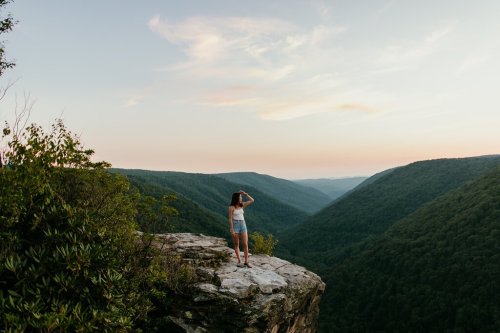 A person stands on a rocky ledge, gazing at a vast green valley under a pastel sky at sunset.