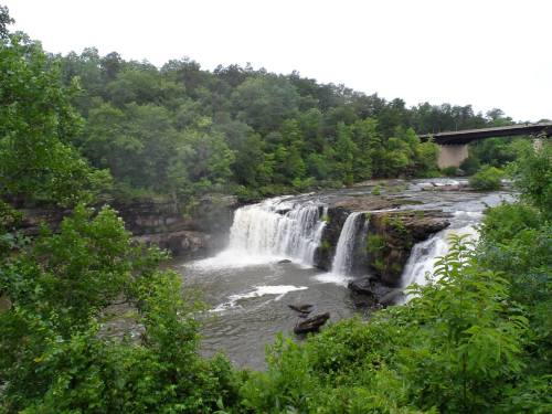 A scenic waterfall surrounded by lush greenery, with a bridge visible in the background.