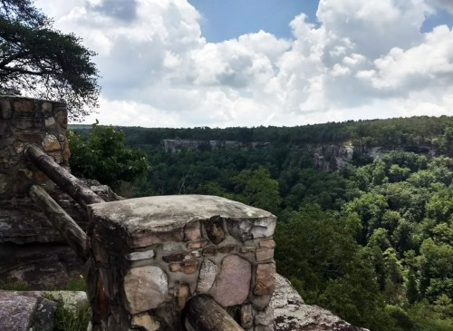 A rocky overlook with a stone railing, showcasing a lush green valley and dramatic clouds in the sky.