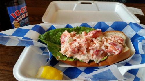 A lobster roll on a bun with lettuce, served in a takeout container, alongside a lemon wedge and a bottle of root beer.