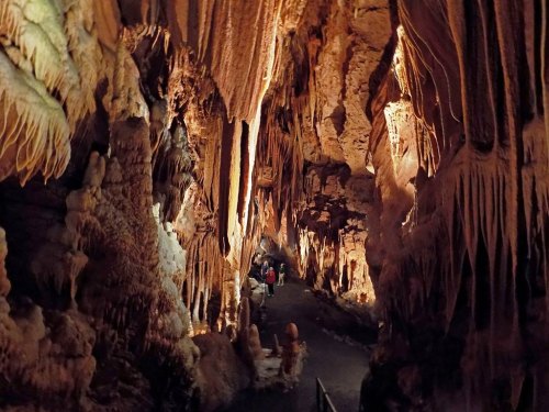 A dimly lit cave with stalactites and stalagmites, featuring a winding path and visitors exploring the underground landscape.