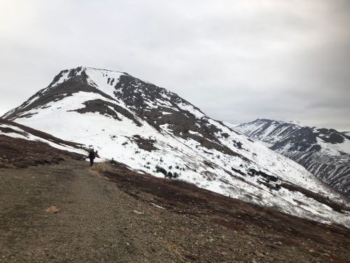 A hiker walks along a rocky path with snow-capped mountains in the background under a cloudy sky.