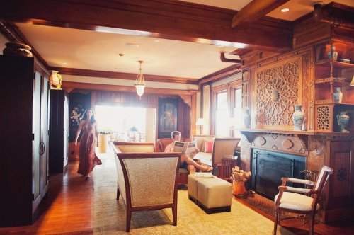 A cozy living room with wooden accents, featuring a woman walking and a man reading a newspaper by the fireplace.