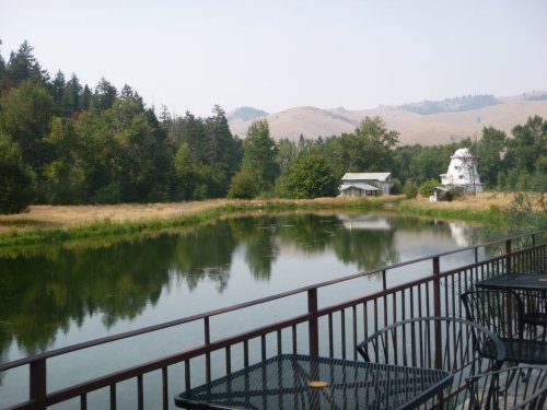A serene landscape featuring a calm pond, surrounded by trees and hills, with outdoor tables in the foreground.