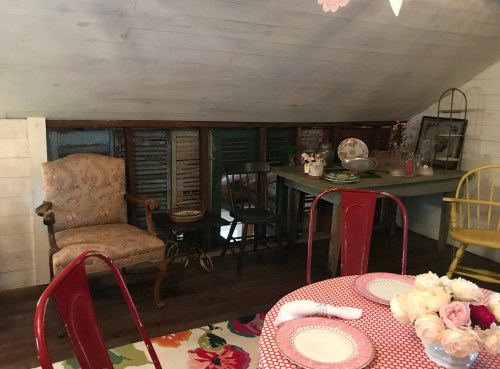 Cozy attic space with vintage chairs, a table, and colorful decor, featuring rustic wooden shutters in the background.