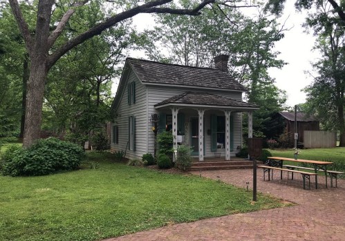A small, charming house surrounded by greenery, with a porch and a picnic table nearby on a brick path.