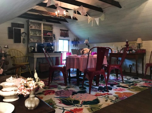 Cozy attic room with a table, red chairs, floral rug, and vintage decor, featuring a warm, inviting atmosphere.