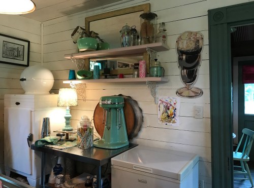 A cozy kitchen corner featuring shelves with vintage decor, a teal mixer, and a lamp, all against wooden walls.