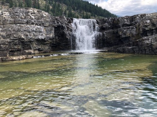 A serene waterfall cascades over rocky cliffs into a clear pool, surrounded by lush greenery and mountains.