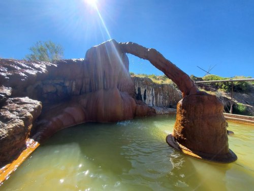 A natural hot spring with a rock arch, surrounded by clear blue skies and lush greenery. Water flows gently into the pool.