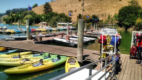 A marina with colorful kayaks, boats, and life jackets, surrounded by greenery and hills on a sunny day.
