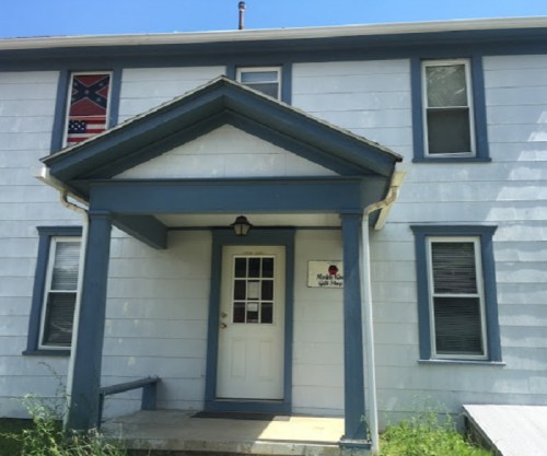 A two-story house with a blue trim, featuring a Confederate flag on the left side and a white door at the entrance.