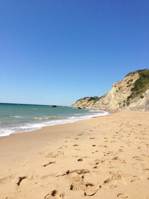 A sandy beach with gentle waves, cliffs in the background, and a clear blue sky. Footprints lead along the shore.