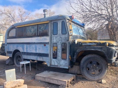 An old, weathered bus with a blue and yellow exterior, parked on uneven ground near trees and scattered debris.