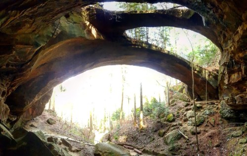 A natural stone archway surrounded by trees, with sunlight streaming through the opening.