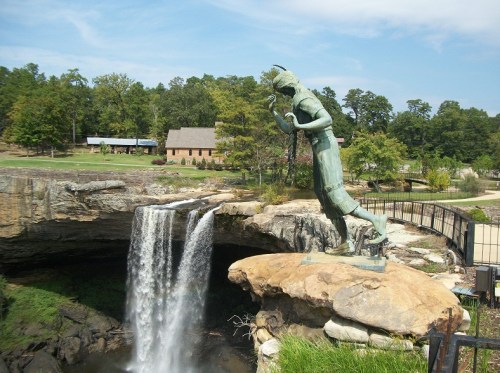 A statue of a Native American figure stands near a waterfall, surrounded by trees and a rustic building in the background.