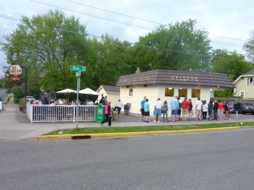 A small ice cream shop with a sign reading "Nelson's," surrounded by trees and a line of customers outside.