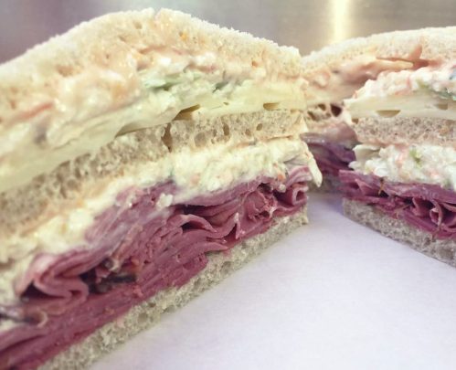 Close-up of a layered sandwich with deli meats, creamy filling, and sliced vegetables between two pieces of bread.