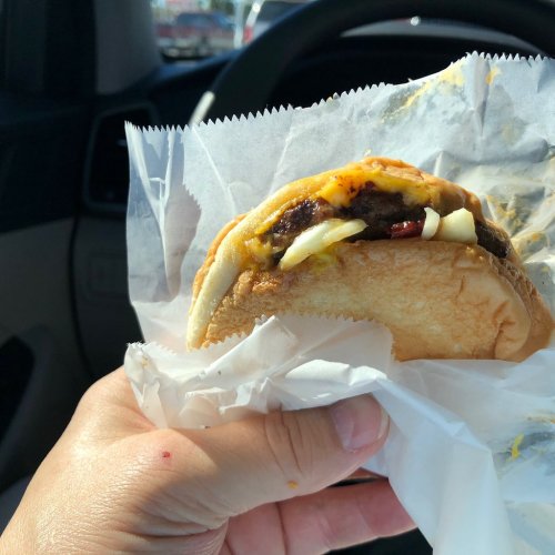 A hand holding a cheeseburger wrapped in paper, with a car interior visible in the background.