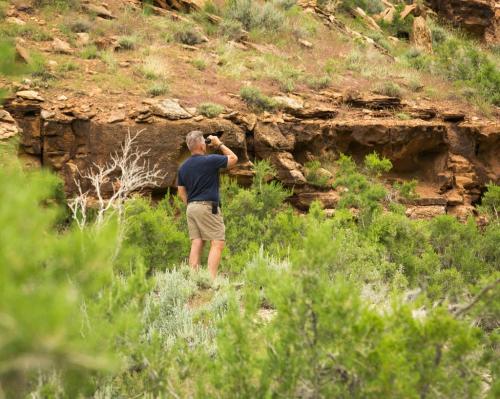 A person stands in a green landscape, photographing rock formations in a rugged, natural setting.