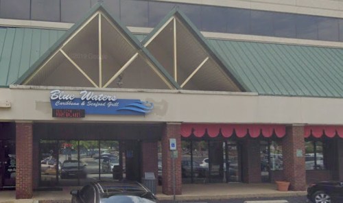 Exterior of Blue Waters Caribbean & Seafood Grill, featuring a green roof and large windows, with parked cars in front.