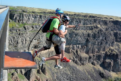 A person in a harness prepares to bungee jump off a cliff with an instructor, overlooking a rocky canyon.