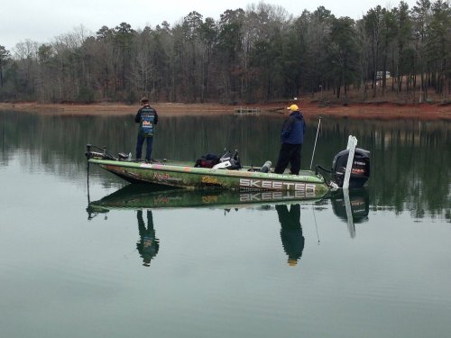 Two fishermen stand on a boat in calm waters, surrounded by trees reflecting on the surface.