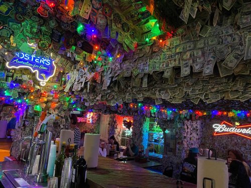 A vibrant bar interior adorned with colorful lights and walls covered in dollar bills and stickers.