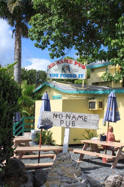 A colorful pub sign reading "No Name Pub" surrounded by palm trees and picnic tables in a tropical setting.