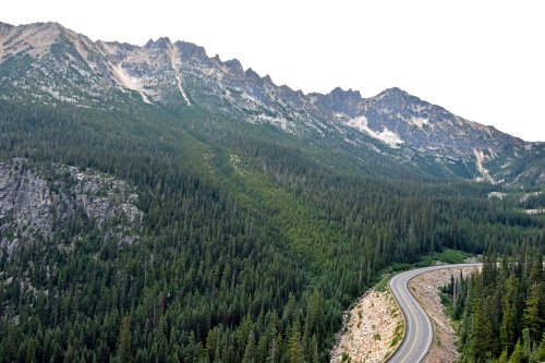 A winding road curves through a dense forest, with rugged mountains rising in the background under a cloudy sky.